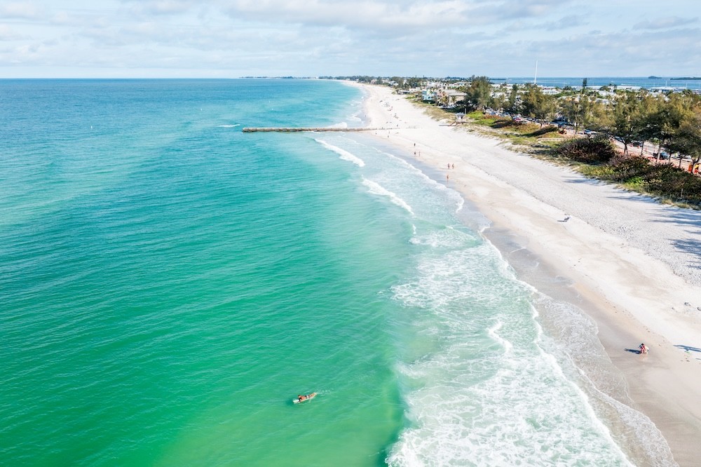 aerial view of Cortez Beach with turquoise water and a bright sunny sky, a surfer is visible as well as a long concrete pier